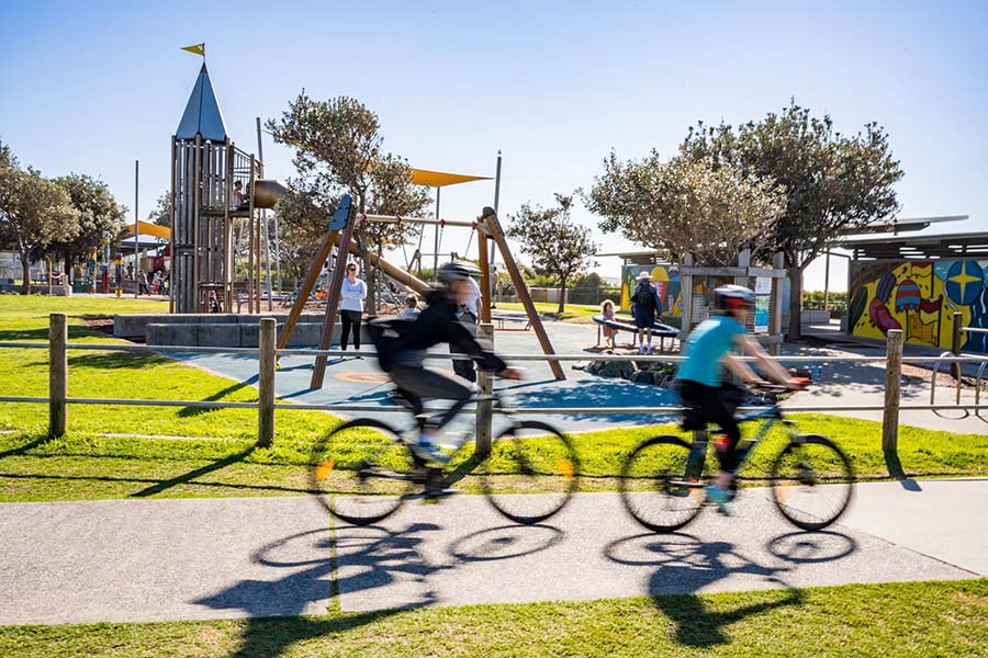 Cyclists ride past Towradgi Beach Park playground, near Wollongong NSW. Credit: NSW Department of Planning, Housing and Infrastructure / Dee Kramer